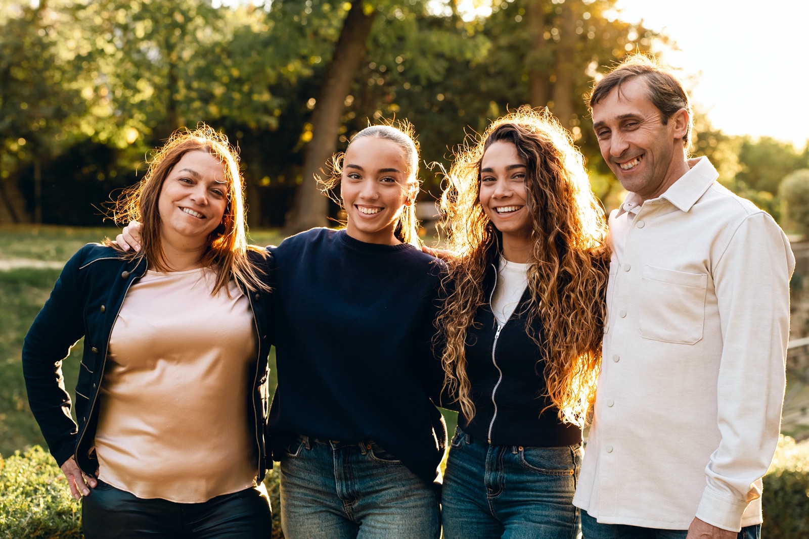 Une famille de quatre personnes souriantes, serrées les unes contre les autres dans un parc, à la lumière du soir.