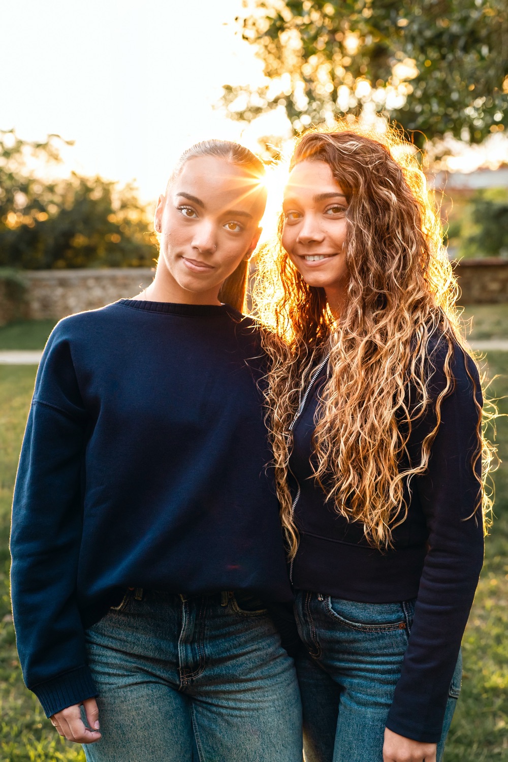 Deux jeunes femmes côte à côte devant le soleil couchant, l'une aux cheveux tirés, l'autre aux longues boucles dorées.