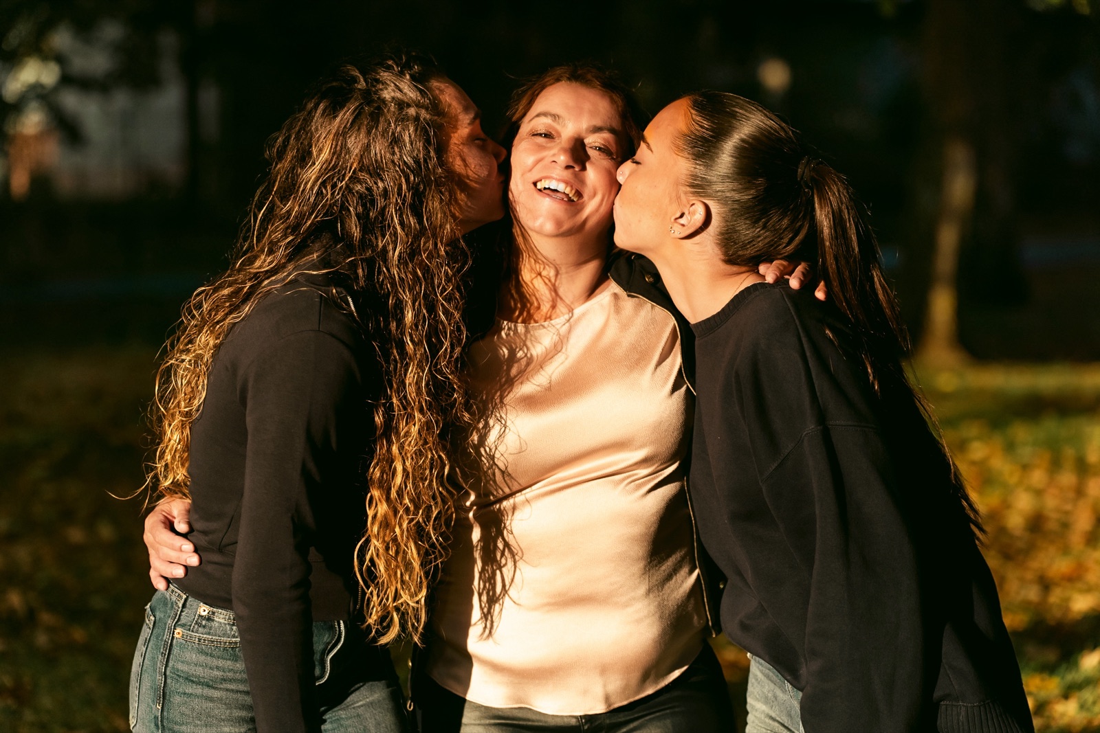 Une femme qui rit, embrassée sur les deux joues par deux jeunes femmes, dans une lumière chaude de fin de journée.