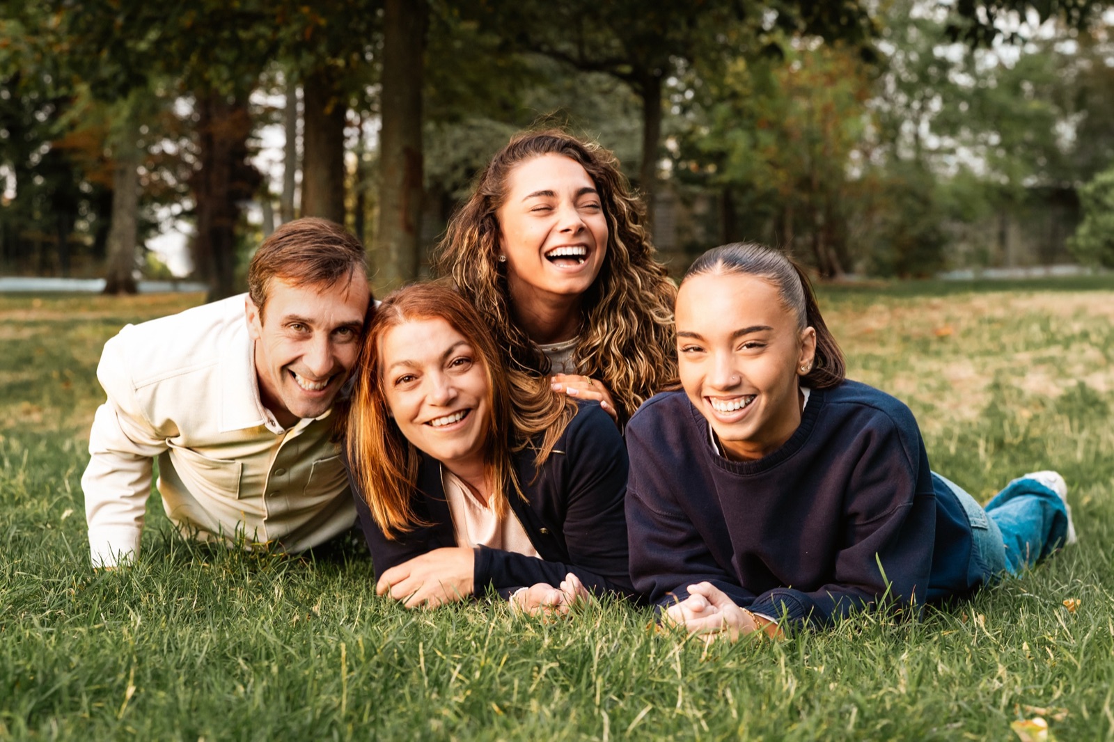 Une famille allongée dans l'herbe d'un parc, souriante et serrée, en lumière douce d'automne.