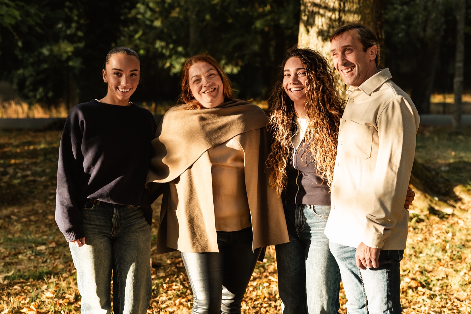 Quatre personnes souriantes dans un parc, debout côte à côte dans la lumière du soir, au milieu des feuilles d'automne.