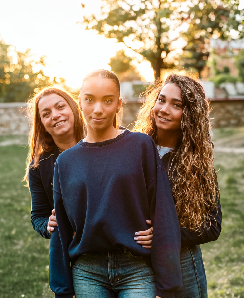 Trois femmes souriantes en lumière de fin de journée : une jeune femme au premier plan, une femme et une autre jeune femme aux longs cheveux bouclés derrière elle.
