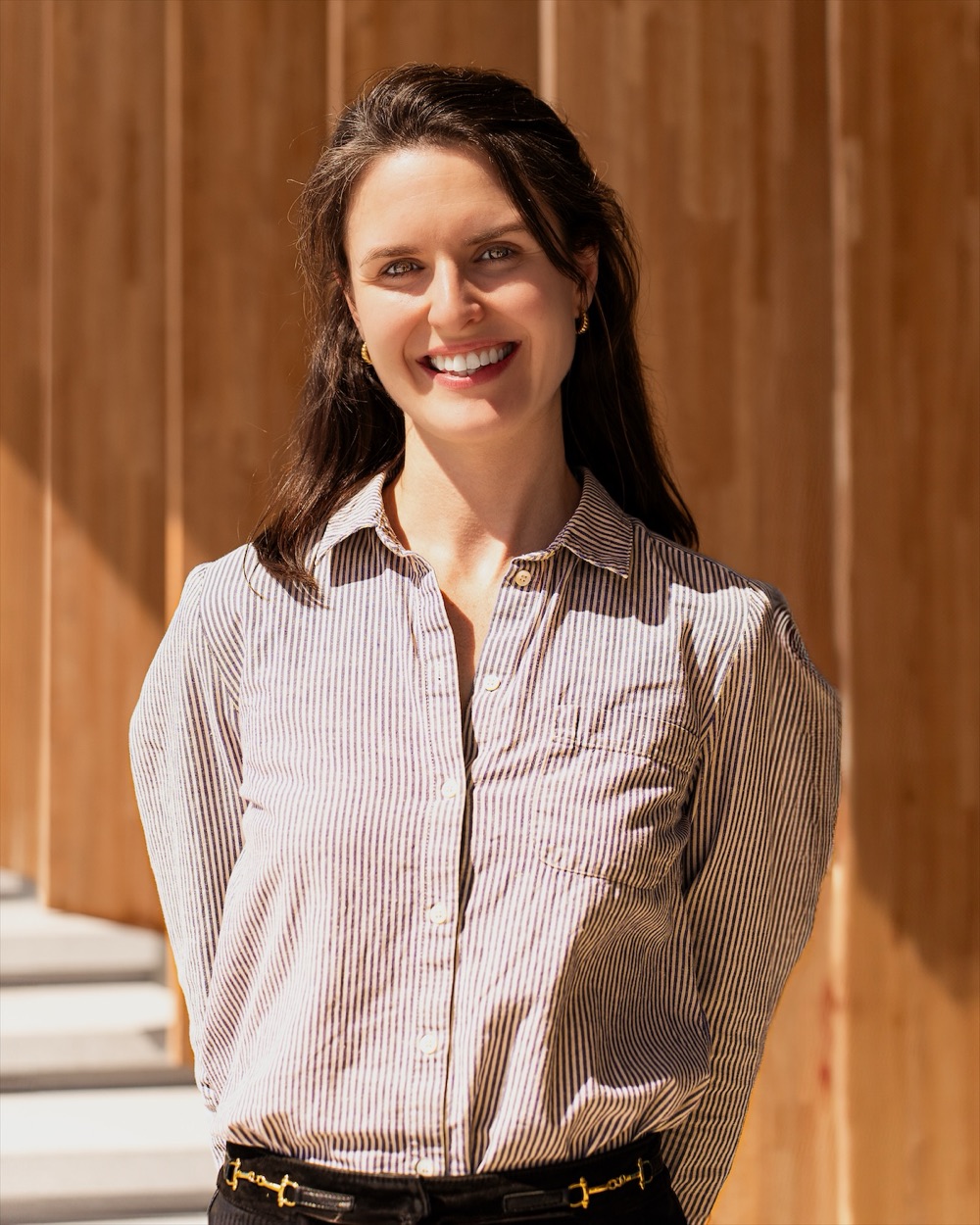 Portrait en extérieur d'une femme souriante en chemise rayée, devant une façade en bois.