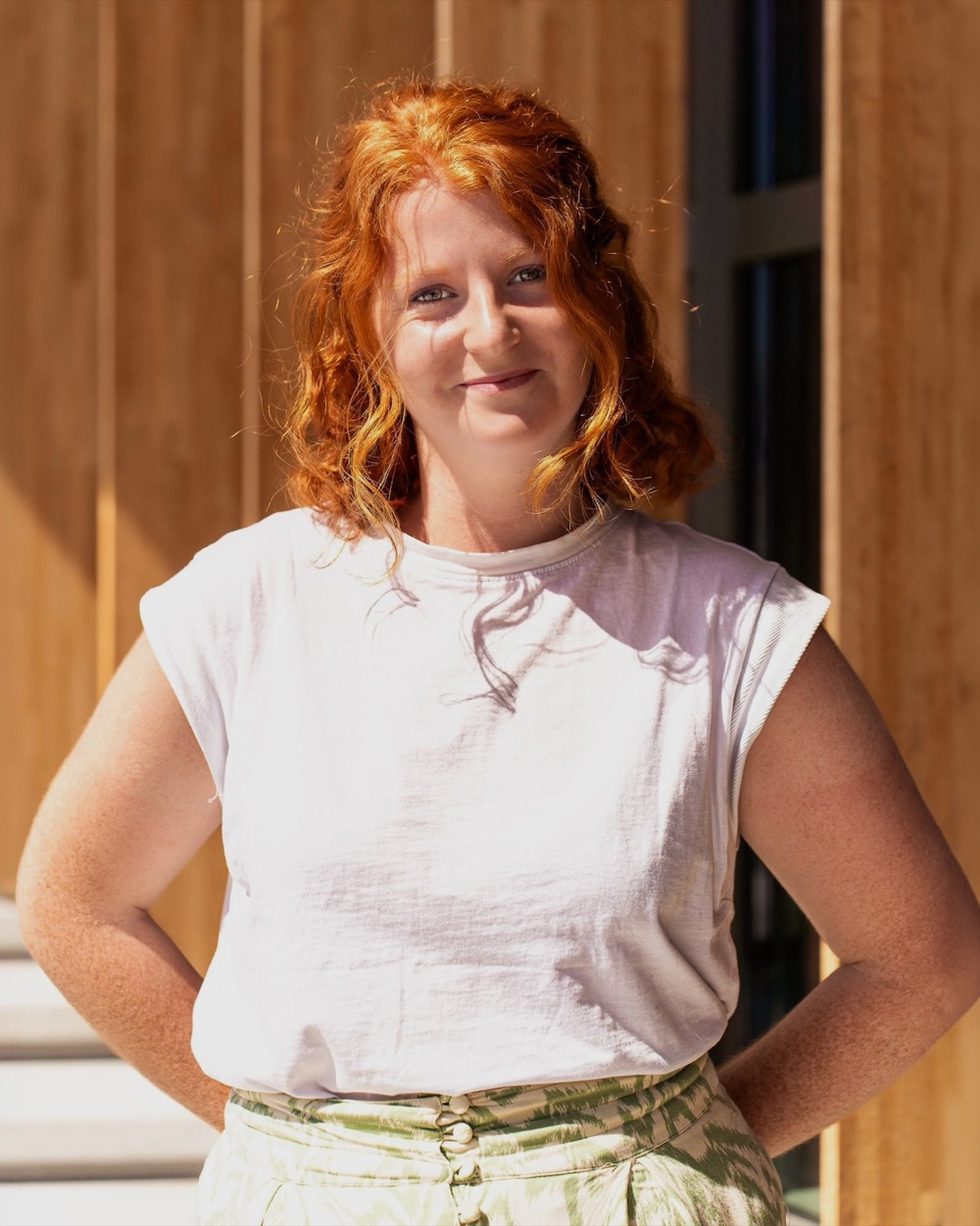 Portrait en extérieur d'une femme rousse souriante, les mains sur les hanches, devant une façade en bois.