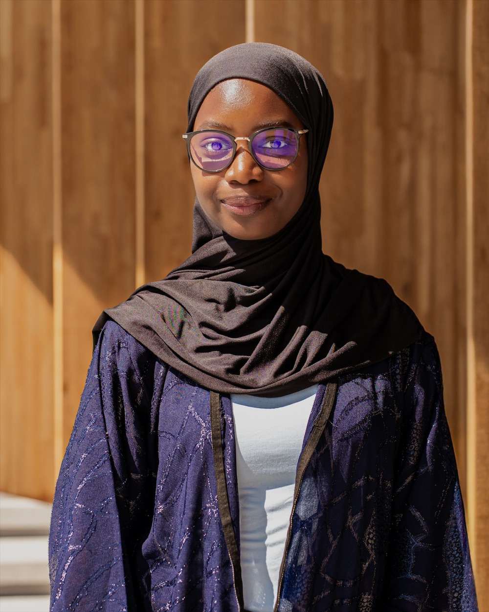 Portrait en extérieur d'une femme à lunettes, regard direct, devant une façade en bois.