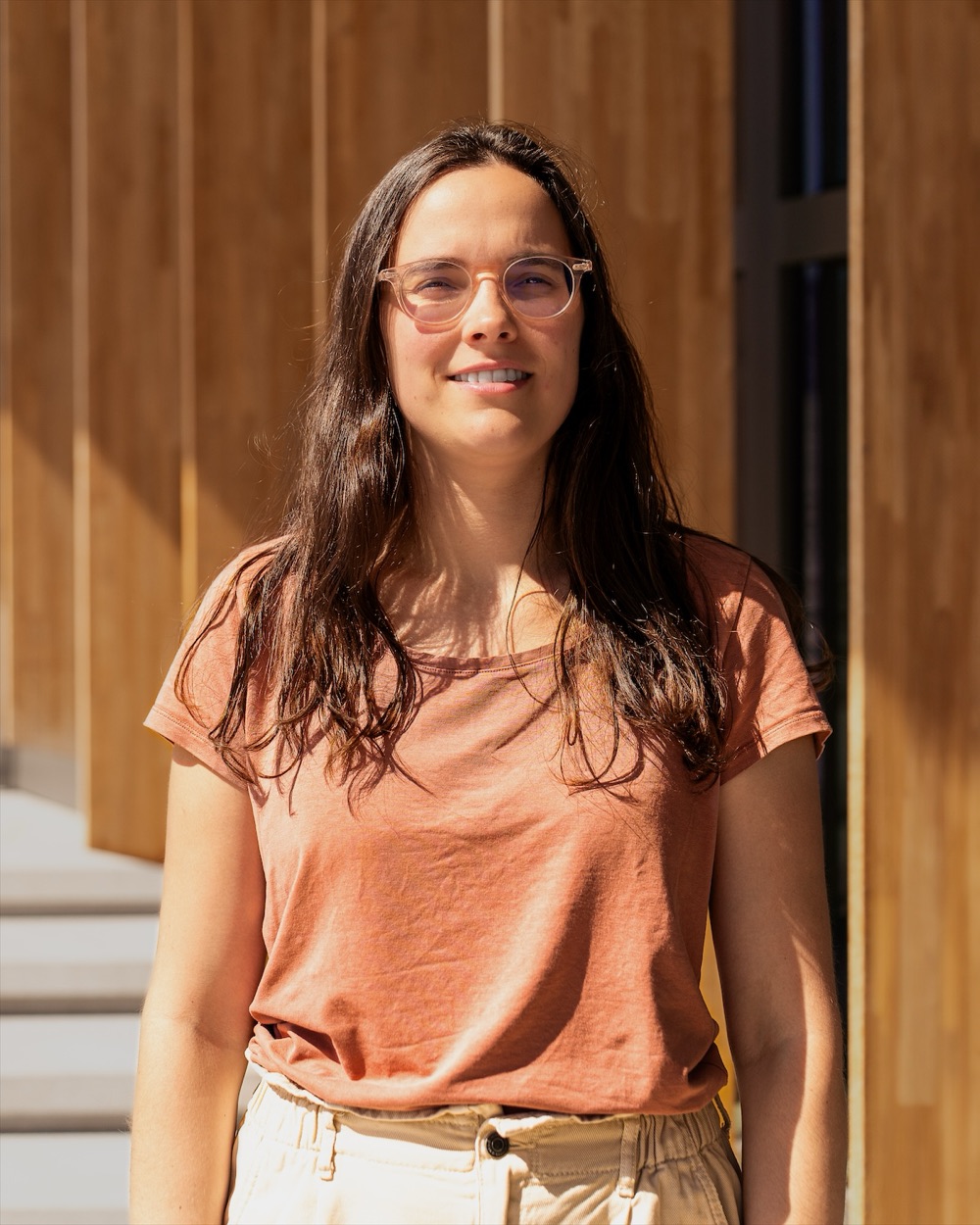 Portrait en extérieur d'une femme à lunettes claires et aux cheveux longs, devant une façade en bois.